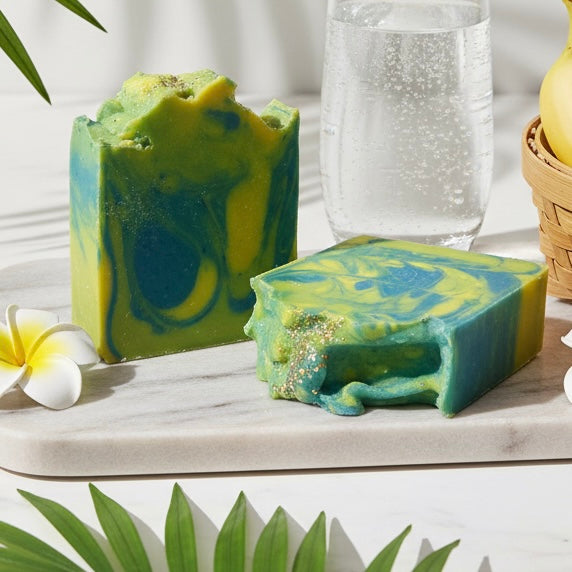 Two green and blue marbled soap bars on a marble surface with a glass of water and flowers in the background.
